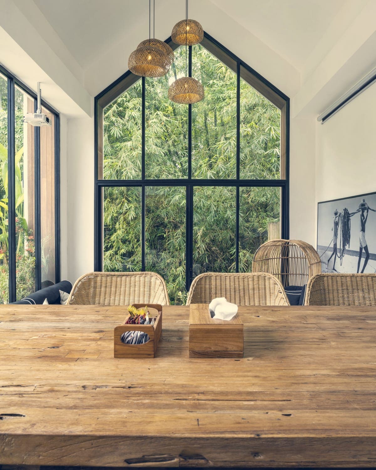 A worn wooden dining table and wicker chairs under a tall gabled window, with green trees outside